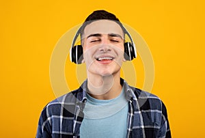 Handsome young man wearing headphones, listening to music with closed eyes on orange studio background