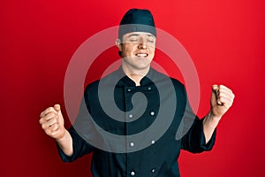 Handsome young man wearing professional cook uniform and hat excited for success with arms raised and eyes closed celebrating