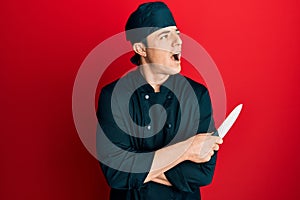 Handsome young man wearing professional cook uniform holding knife angry and mad screaming frustrated and furious, shouting with