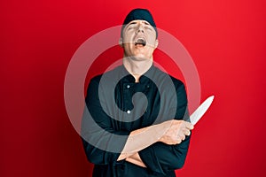 Handsome young man wearing professional cook uniform holding knife angry and mad screaming frustrated and furious, shouting with