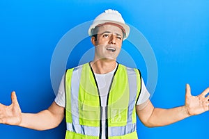 Handsome young man wearing safety helmet and reflective jacket crazy and mad shouting and yelling with aggressive expression and