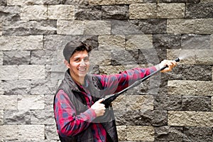 Handsome young man wearing square pattern red holding high pressure water gun, pointing towards grey brick wall photo