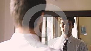 Handsome young man in white shirt stands by the mirror wearing a tie