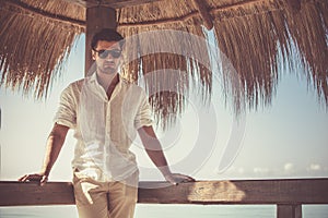 Handsome young man with white shirt and sunglasses relaxed leaning on a wooden bar. During the summer, with reed leaves in the