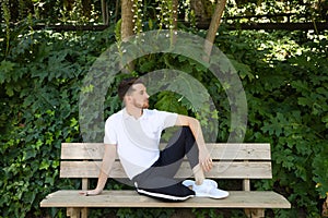 Handsome young man in white T-shirt and black trousers sitting on the wooden bench in the park waiting impatiently for someone to