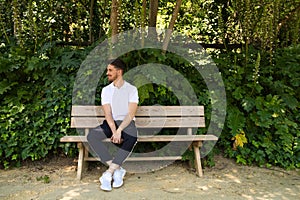 Handsome young man in white T-shirt and black trousers sitting on the wooden bench in the park waiting impatiently for someone to