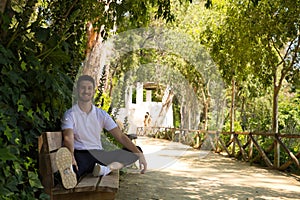 Handsome young man in white T-shirt and black trousers sitting on the wooden bench in the park waiting impatiently for someone to