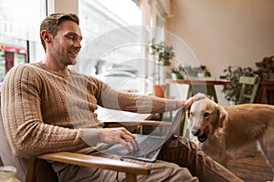 Handsome young man working in cafe with a dog, sitting on chair and using laptop, petting his golden retriever in animal