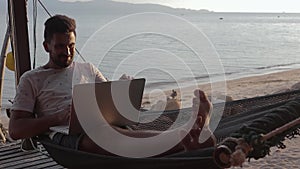 A handsome young man working on his laptop in the hammock on the beach.