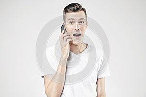 Handsome young man yelling with open mouth talking by mobile phone, isolated on white background, wearing white t-shirt.