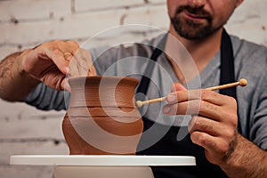 Handsome young potter man in a gray sweater and a black apron sculpts clay pot on a potter`s wheel in a cozy craft