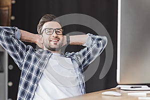 handsome young programmer in eyeglasses smiling at camera while sitting with hands behind head