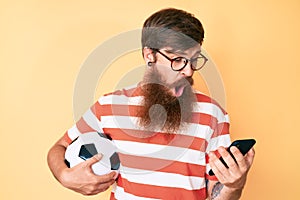 Handsome young red head man with long beard holding football ball looking at smartphone afraid and shocked with surprise and