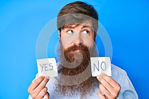 Handsome young red head man with long beard holding yes and no reminder smiling looking to the side and staring away thinking