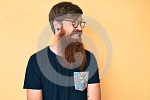 Handsome young red head man with long beard wearing casual clothes and glasses looking away to side with smile on face, natural