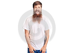 Handsome young red head man with long beard wearing casual white tshirt sticking tongue out happy with funny expression