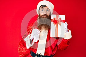 Handsome young red head man with long beard wearing santa claus costume and big bag with presents skeptic and nervous, frowning