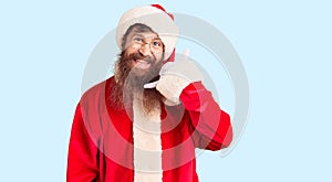 Handsome young red head man with long beard wearing santa claus costume smiling doing phone gesture with hand and fingers like