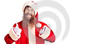 Handsome young red head man with long beard wearing santa claus costume success sign doing positive gesture with hand, thumbs up