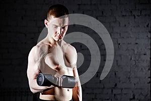 Handsome young shirtless man with muscular body doing exercises using dumbbell against a brick wall.