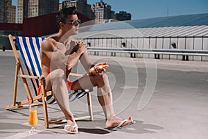 handsome young shirtless man sitting on sun lounger and applying sunscreen lotion