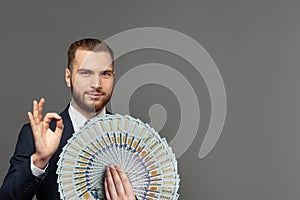 Handsome young smart businessman showing OK gesture with us dollars banknotes fan on grey wall background