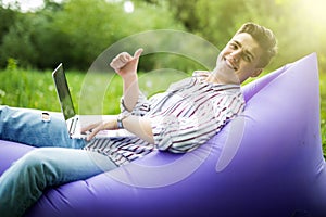 Handsome young smile man lying on inflatable sofa lamzac working on laptop and gesturing thumbs up while resting on grass in park