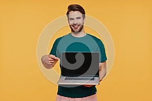 Handsome young smiling man in casual clothing carrying laptop and pointing monitor