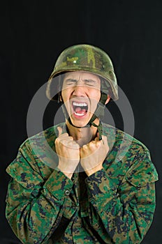 Handsome young soldier wearing uniform suffering from stress, screaming with both hands doing fist, in a black