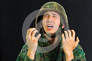 Handsome young soldier wearing uniform suffering from stress, screaming with both hands open, in a black background