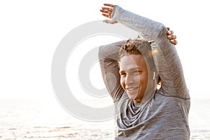 Handsome young sports man standing on the beach listening music with earphones make stretching exercises.