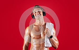 Handsome young sportsman with bare torso and towel listening to music in headphones on red studio background