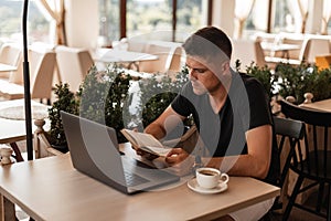 Handsome young student man in a trendy black T-shirt with a laptop with a cup of coffee sitting in a cafe is reading a book.