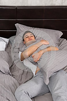 Handsome young tired man in pajama sleeping alone without blanket and hugging a pillow in stylish bed
