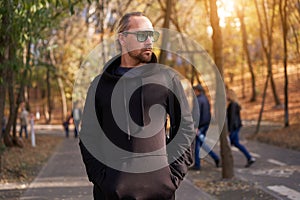 Handsome young unshaven man black hoodie and sunglasses walking autumn park warm day