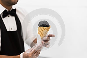 Handsome waiter in elegant classic suit serving trendy dessert, black ice cream in waffle cone. Uninsulated white