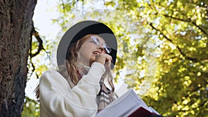 Handsome young woman in the stylish sunglasses writing some thought in her dairy while sit in the green park.