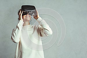 Handsome young woman wearing virtual headset. Smiling hipster using VR glasses. Blank sweater. Grey concrete wall background