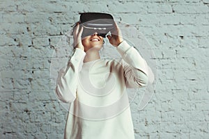 Handsome young woman wearing virtual headset. Smiling hipster using VR glasses. Blank sweater. White bricks wall background