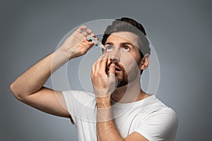 Handsone bearded caucasian man applying ophthalmology eyedropper, standing on grey studio background