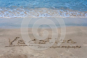Handwriting the words `Vacation` on sand beach with white waves of blue sea in the background.