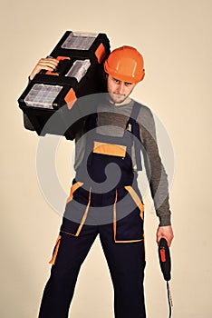 Handyman concept. Man in overall and helmet holds toolbox on shoulder and drill in hand. Strong and attractive repairer
