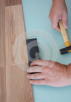 A handyman is installing a wood laminate flooring on the underlayment using a hummer along with a tapping block to snug up the joi