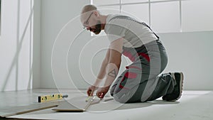 Handyman in jumpsuit measures part of parquet plank on knees