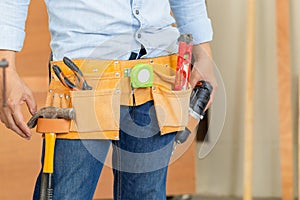 Handyman manual worker in tools belt and holding drill in his hands, carpenter working with equipment in wood workshop, man doing