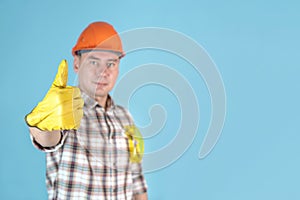 A handyman in an orange helmet and gloves shows a thumbs up standing on a blue background. Copyspace on a blue background,