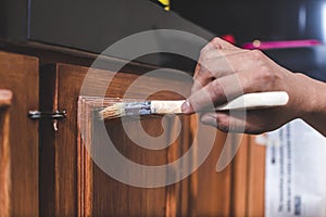 A handyman paints a fresh coat of varnish on the surface of a base kitchen cabinet with a medium sized brush. Home renovation or