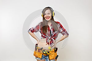 Handyman woman in shirt, denim shorts, noise insulated headphones, kit tools belt full of instruments standing with arms