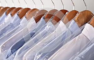 Hangers with shirts in dry cleaning plastic bags on rack against light background, closeup