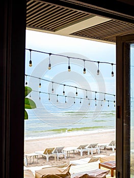 Hanging festive string wired light bulbs decorate under the roof outside the cafe on beachfront with seascape view background.